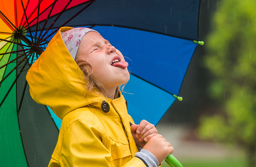 Girl in a yellow rain jacket holding a rainbow umbrella trying to catch a raindrop on her tongue